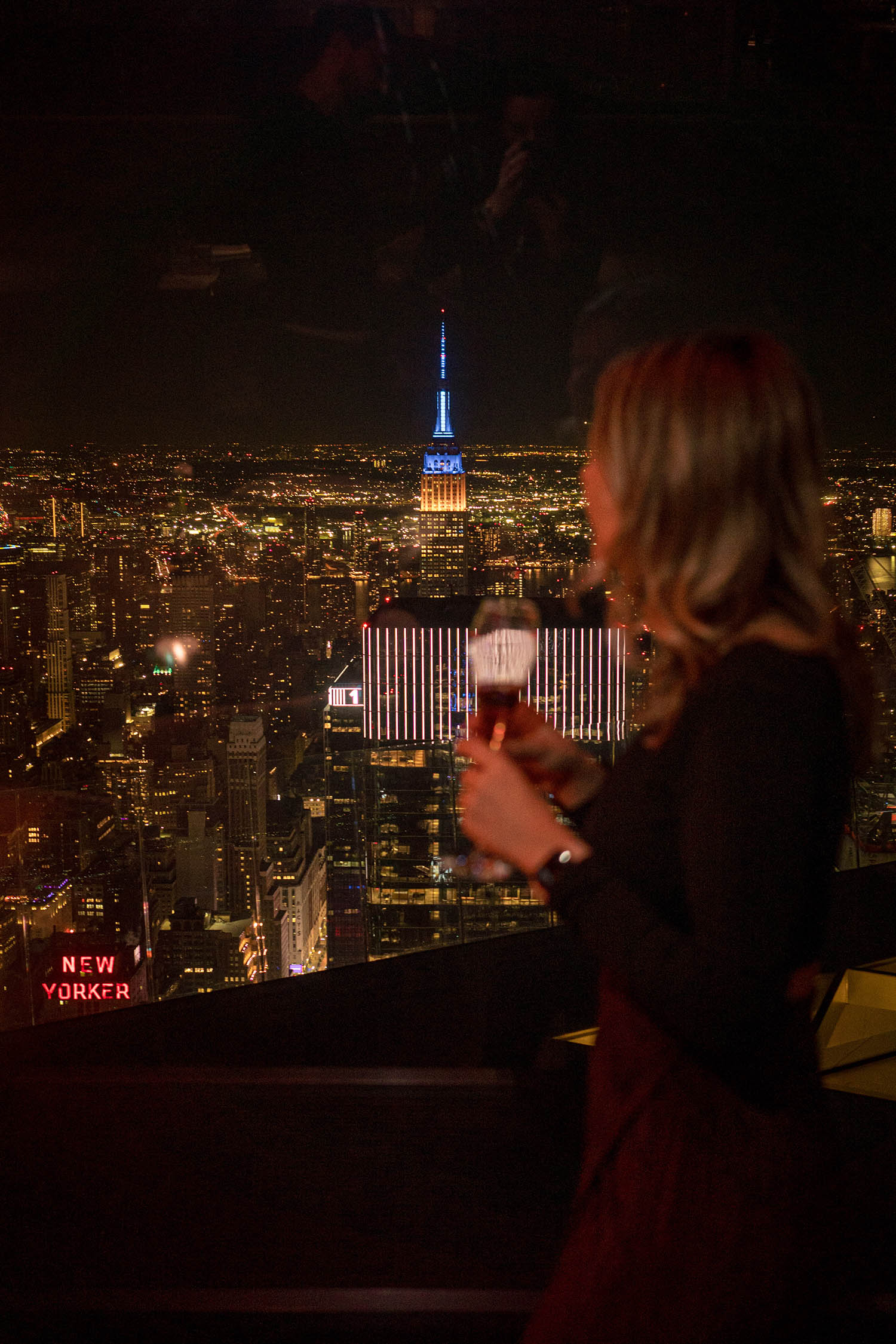 Silhueta de uma mulher no restaurante Peak, com taça de bebida e o Empire State ao fundo.